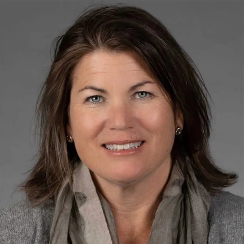 Smiling woman with shoulder-length brown hair wearing a gray scarf and cardigan against a gray background.