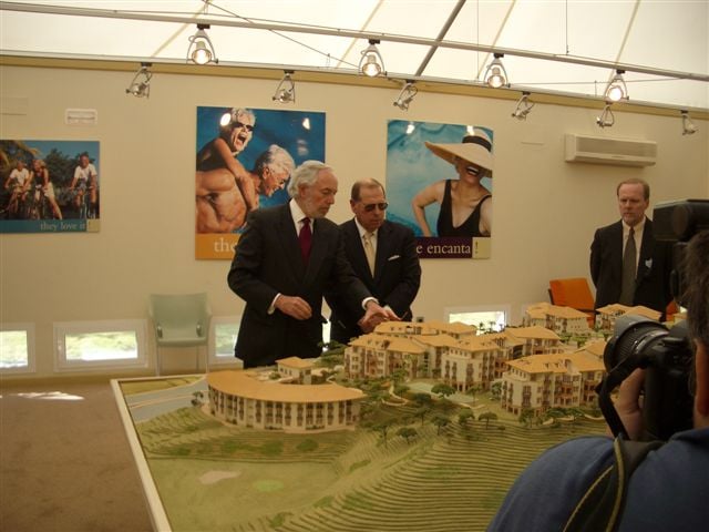 Two suited men present a detailed resort scale model while cameras record in a brightly lit showroom.