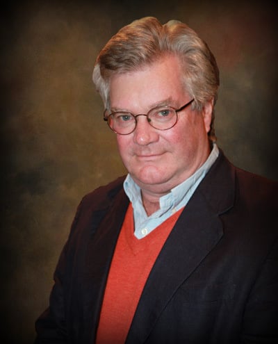 Studio portrait of a gray-haired man wearing glasses, a dark blazer, red sweater, and light collared shirt.