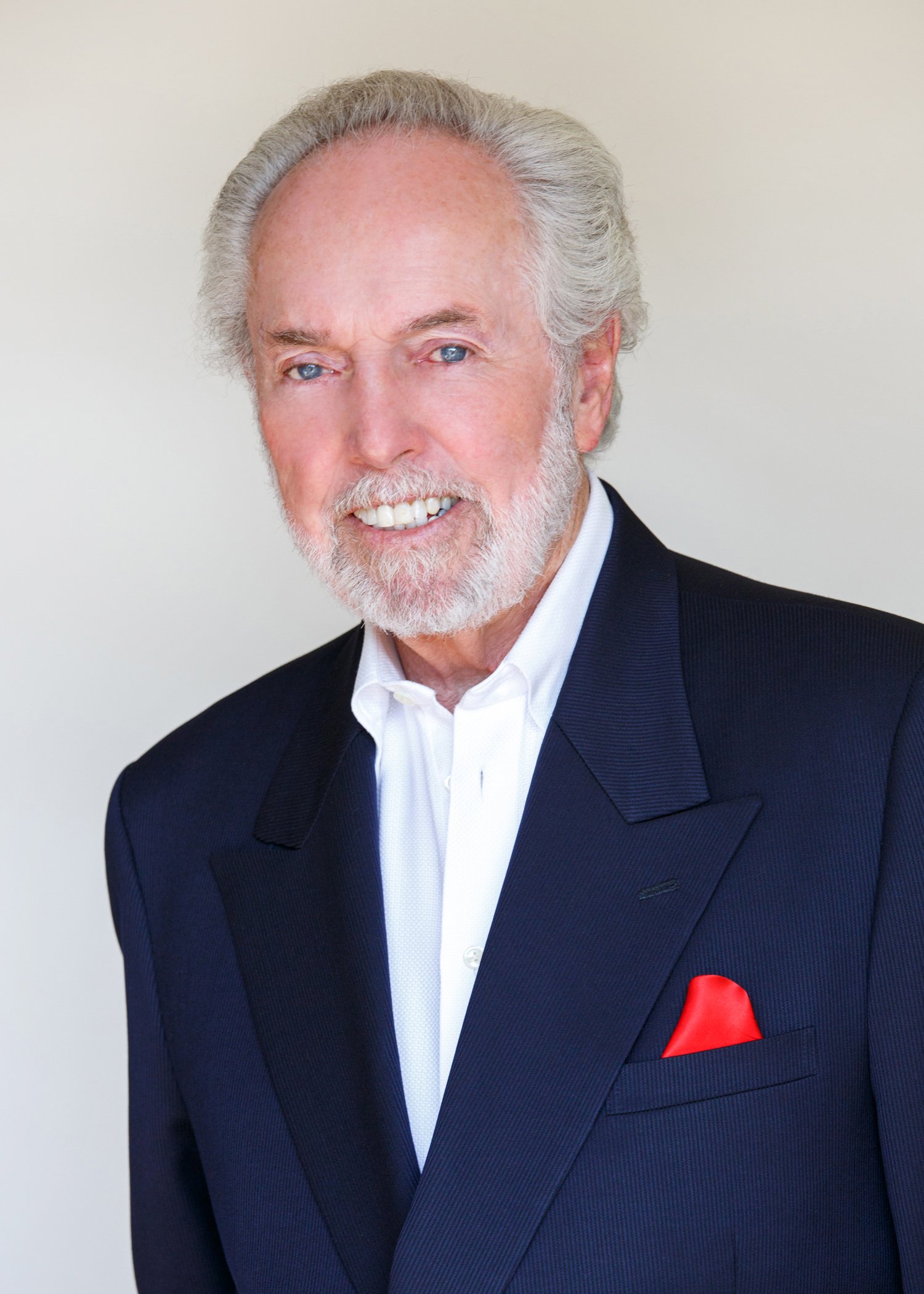 Smiling older man with gray hair and beard wearing a navy pinstriped suit and red pocket square.